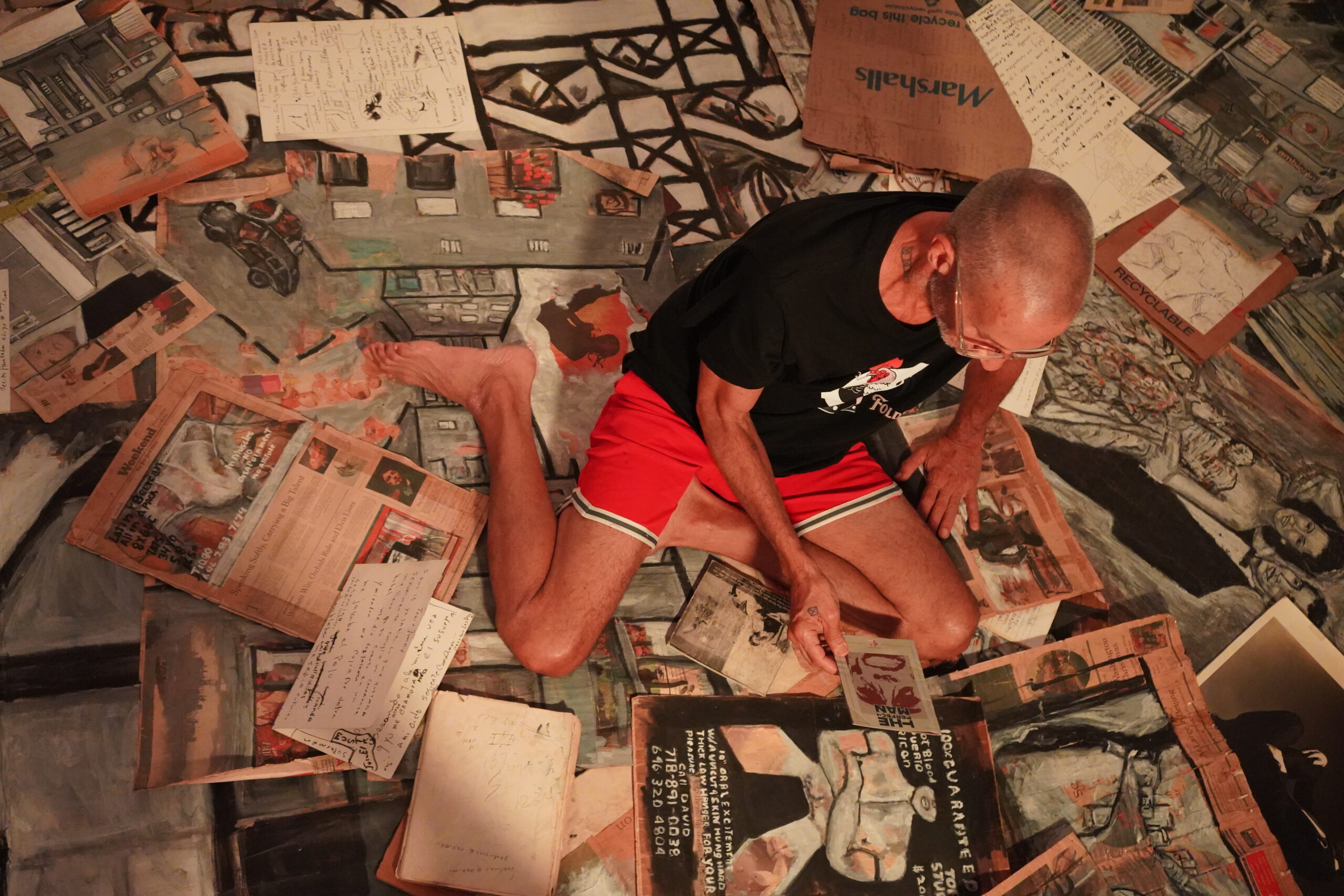 A person in red basketball shorts and a black shirt sits on a floor covered with scattered artwork and documents, including sketches and pages with text, examining a piece of paper