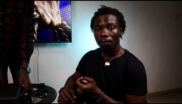 A person with dark skin and a slight mustache looks directly at the camera as another person mostly off camera handles recording equipment