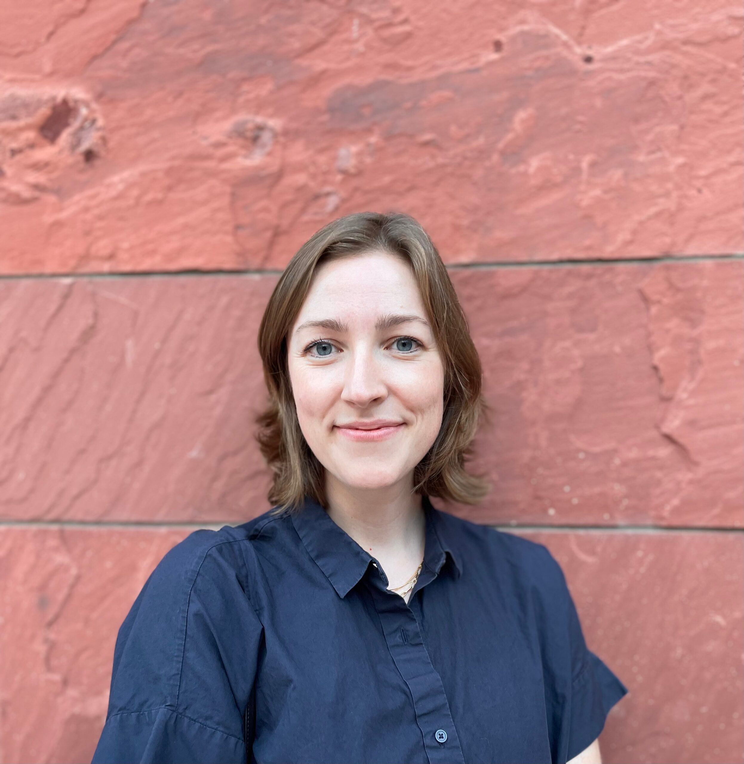A female-presenting person with shoulder length light brown hair and light skin poses in a black collared shirt against a pink textured painting
