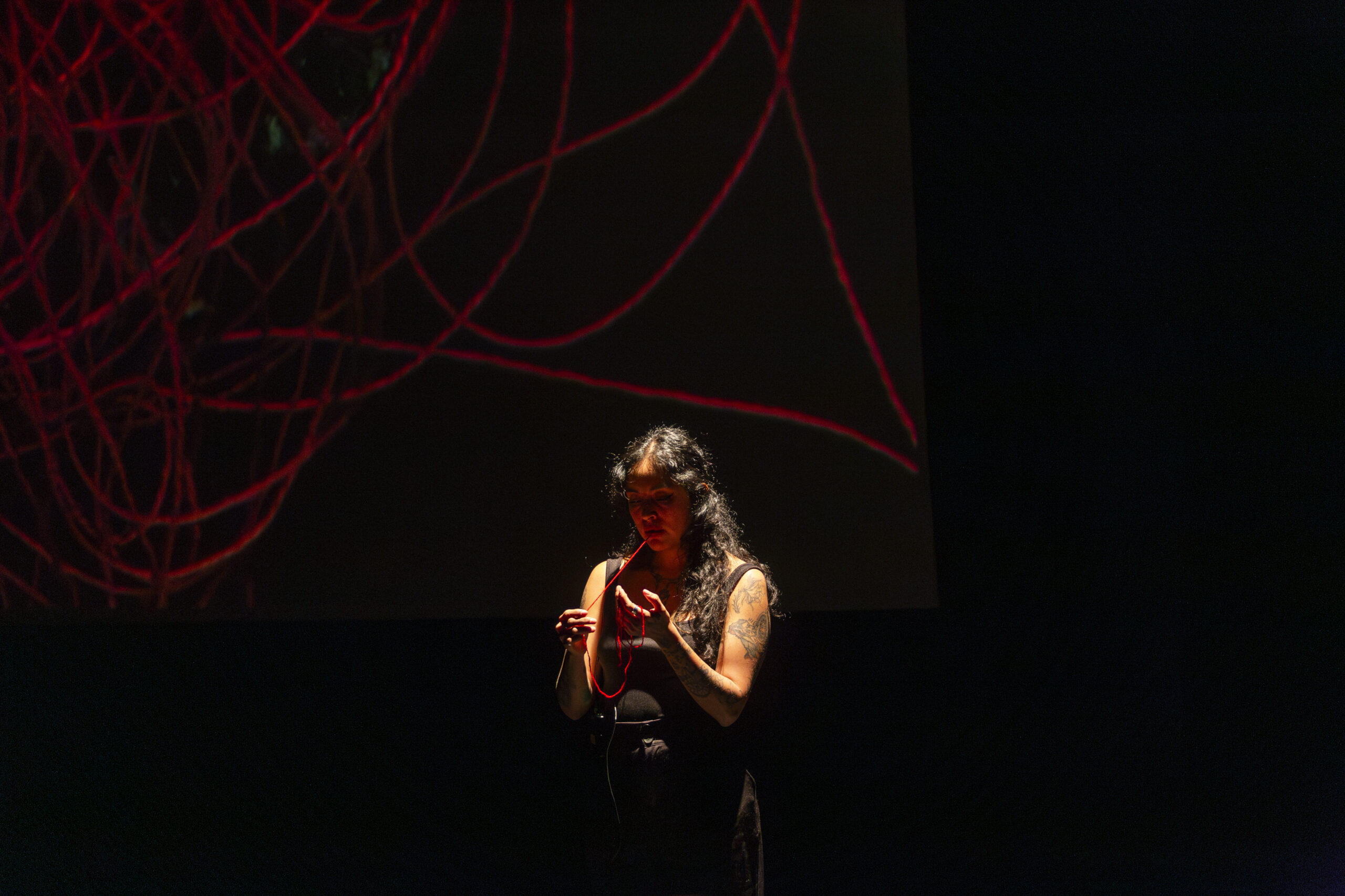 A person with long curly hair looks at their hands while pulling red thread from their mouth in a theater with a black backdrop filled with red scribbles