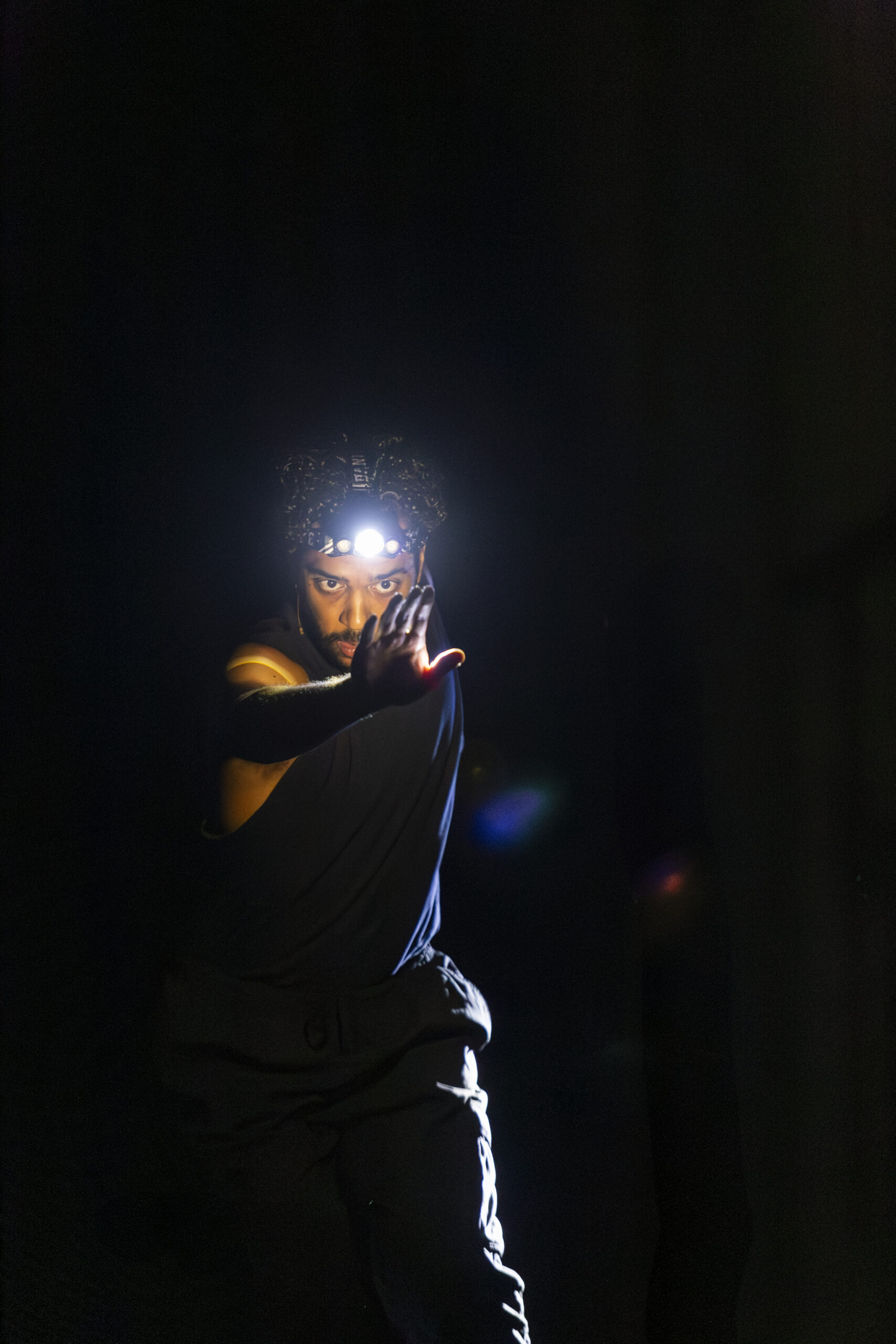 A person dressed in black with a headlight on looks at their right hand against a black background