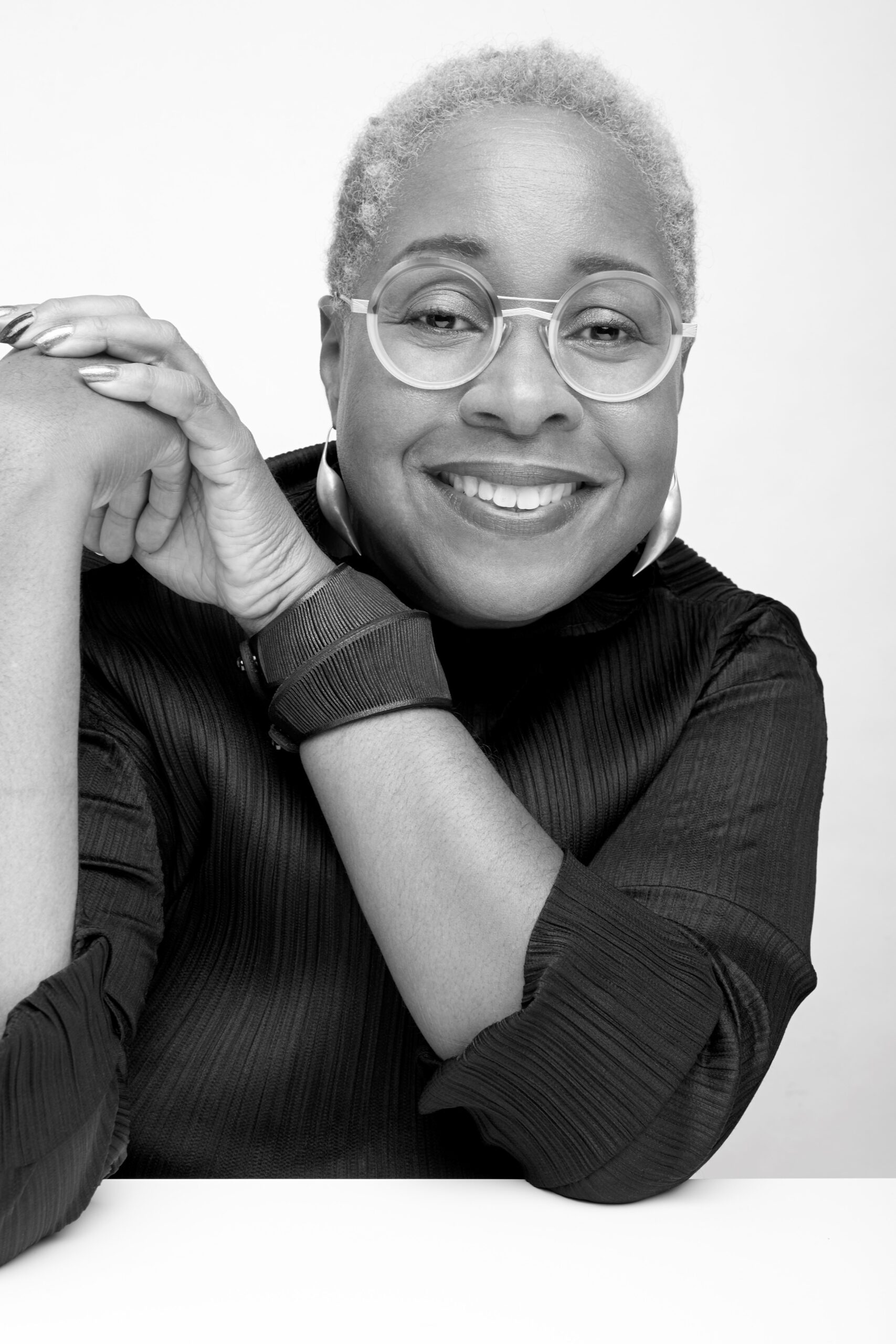 Black and white portrait of a female-presenting person with short hair and glasses posed with their hands clasped and held up by their right ear