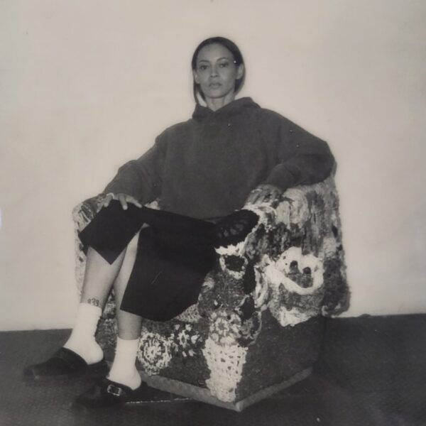 Black and white photo of a female-presenting person dressed in a hooded sweatshirt and skirt sitting in an armchair covered in knitted doilies