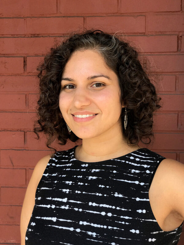 Portrait of a female-presenting person with curly brown hair to their cheekbones and a black tank top with partially complete white stripes posing against a red brick wall