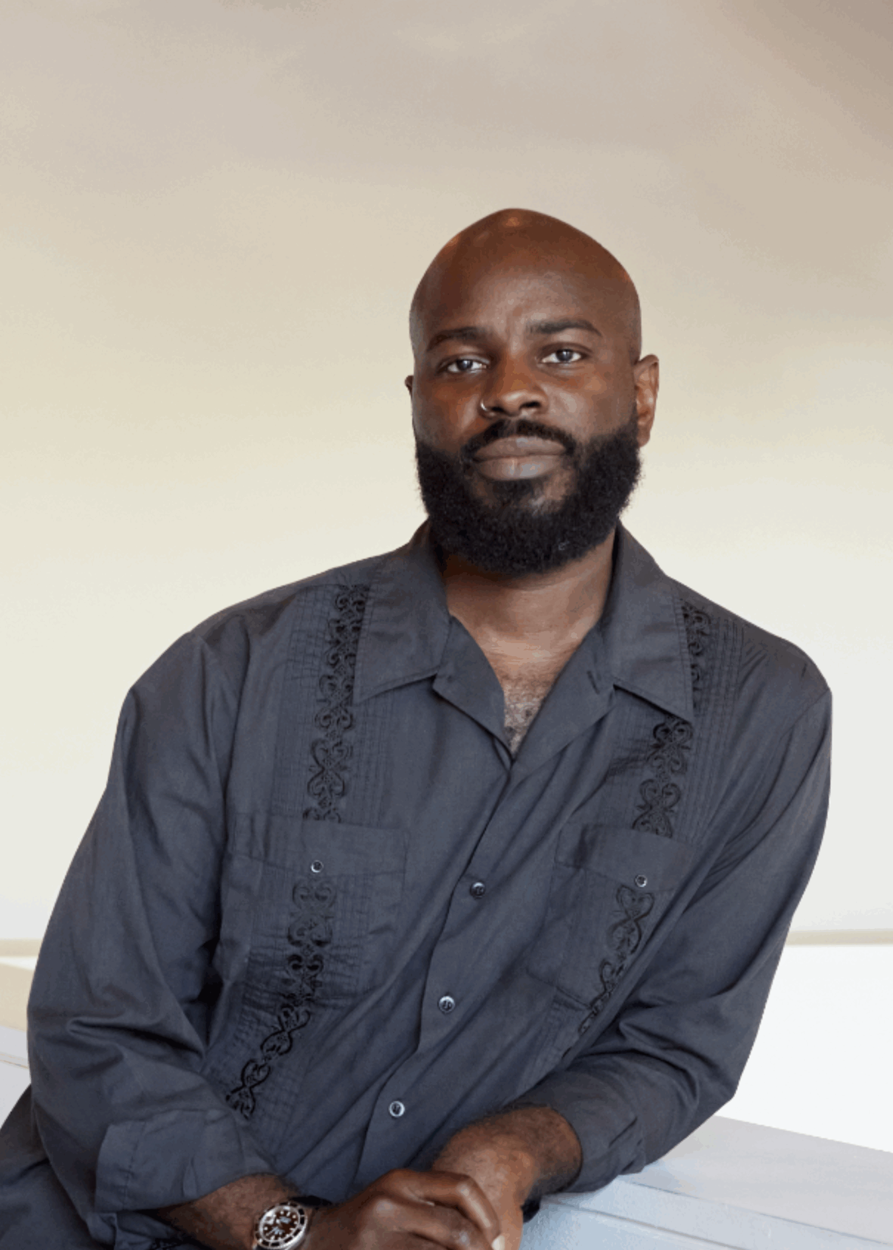 Portrait of a male-presenting person with dark skin and a black beard dressed in a grey-blue shirt and posed against a beige wall