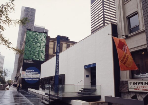 A white two story building with a thin vertical sign reading Museum of Contemporary Art Chicago with skyscrapers behind it