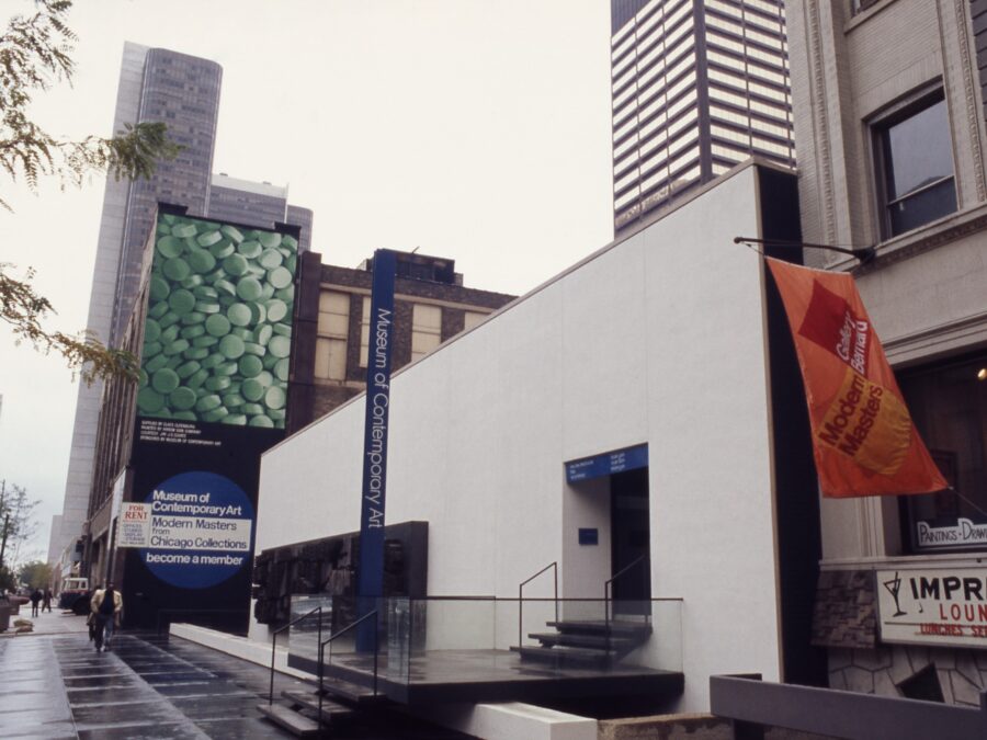 A white two story building with a thin vertical sign reading Museum of Contemporary Art Chicago with skyscrapers behind it