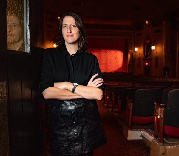 Portrait of a light skinned female presenting person folding their arms and wearing all black in a theater setting