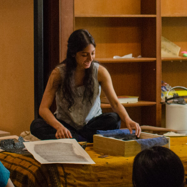 A female presenting person with medium skin and long brown hair sits on a blanket with boxes and artworks in front of them