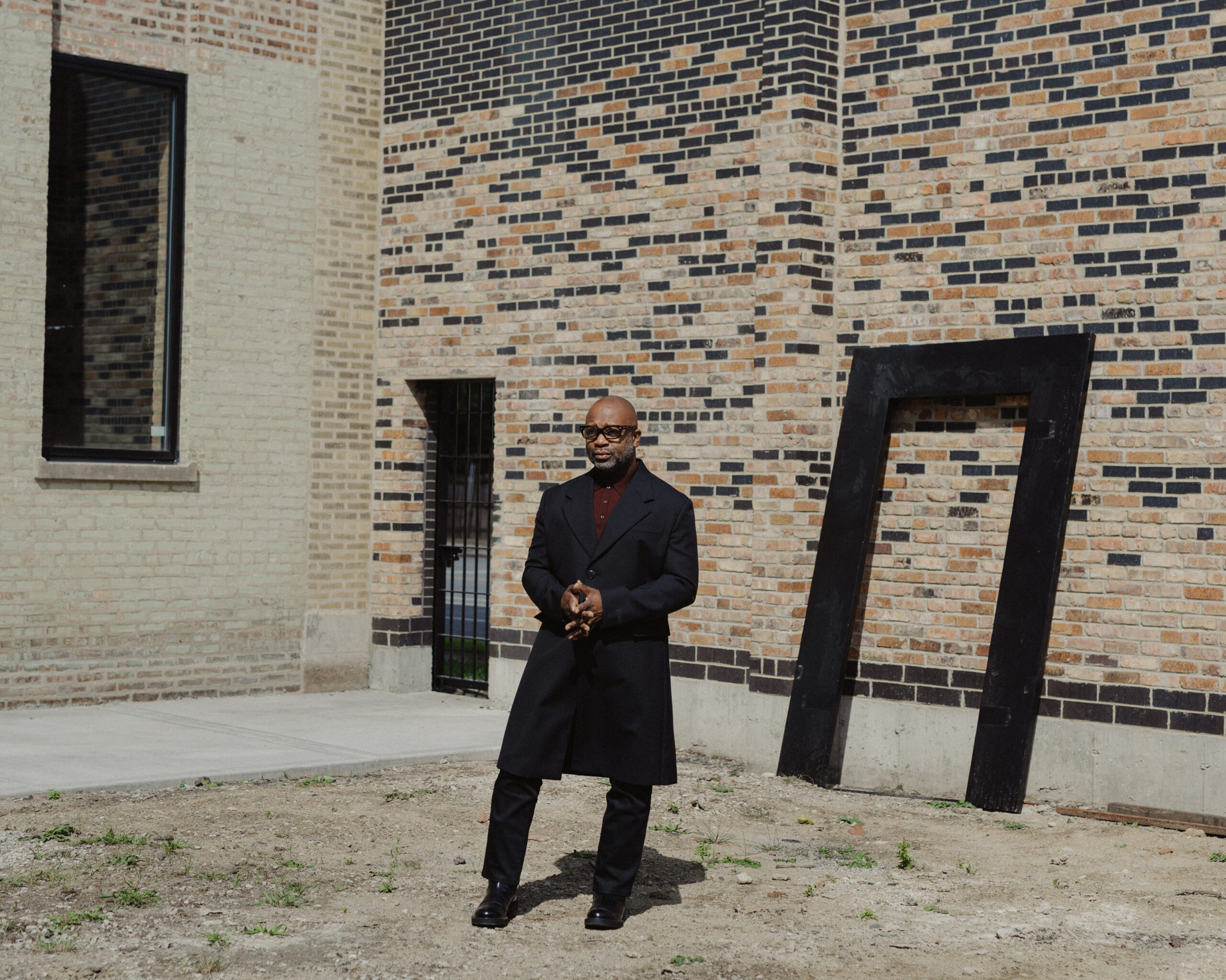A male-presenting person with black glasses dressed in a long black suit coat and pants stands in a courtyard between two brick buildings. A black door frame leans against the building to the right.