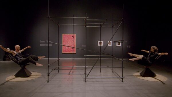 Two people dressed in all black tilt back in black balance chairs in a gallery space painted gray. Between them is a large wire-frame artwork.
