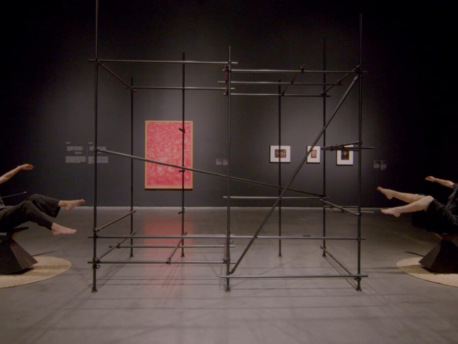 Two people dressed in all black tilt back in black balance chairs in a gallery space painted gray. Between them is a large wire-frame artwork.
