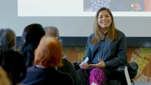 A person with long, light-brown hair sits in front of an audience gesturing with their hands. Magenta yarn is wrapped around their leg.