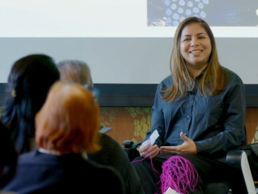 A person with long, light-brown hair sits in front of an audience gesturing with their hands. Magenta yarn is wrapped around their leg.