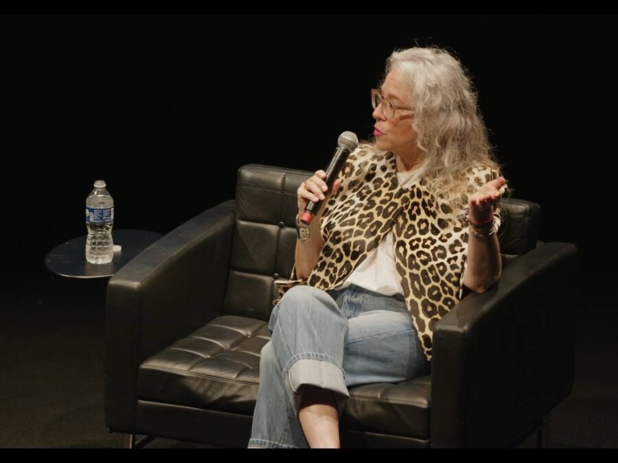 A female-presenting person with light skin dressed in a leopard print shirt and jeans sits on a black stage in a black chair holding a microphone. A small table next to them has a water bottle on it.