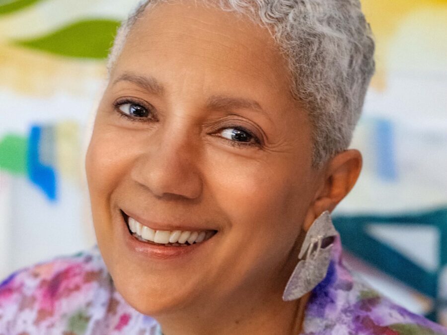 Close up of a female presenting person with short white hair and medium-light skin tilts their head slightly to their left while smiling directly at the camera