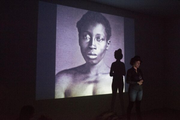 A person stands in front of a large projection screen displaying an archival image of a person with dark skin in a darkened room