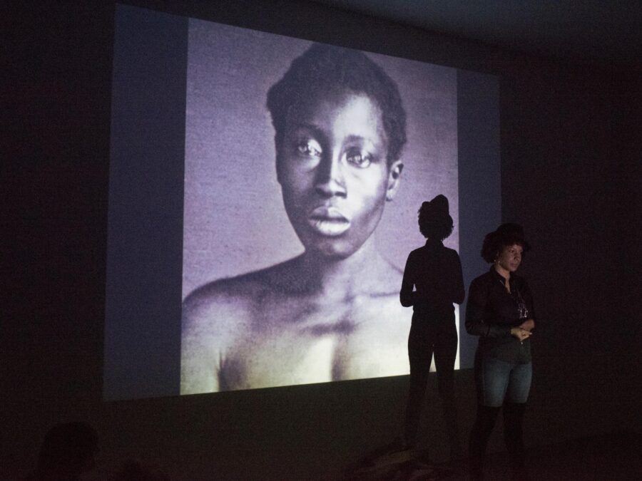 A person stands in front of a large projection screen displaying an archival image of a person with dark skin in a darkened room