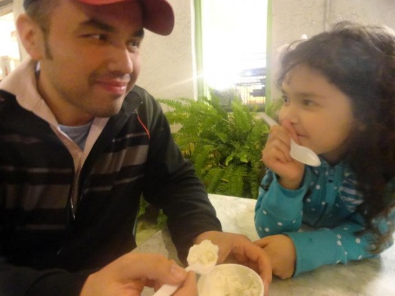 An adult and a child smile at each other at a table in a bright room with plants