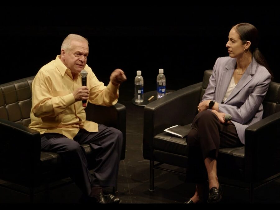 Two people sit in black leather chairs on an all-black stage. A male-presenting person with thinning white hair gestures with their arms towards the female-presenting person with long, dark hair sitting on their left.