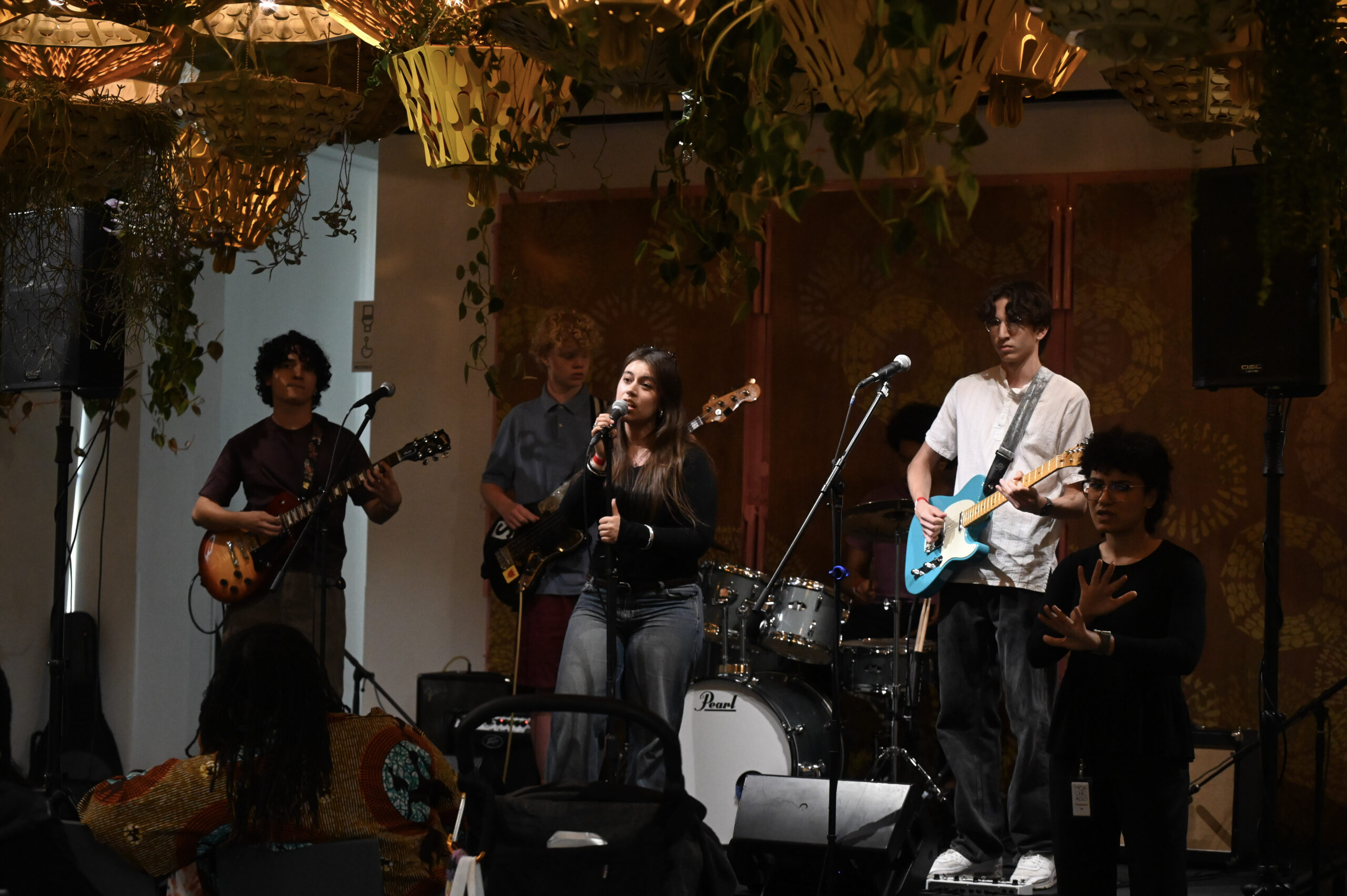 A band performs on stage in a venue decorated with hanging plants. The band consists of a vocalist, two guitarists, a bassist, and a drummer.