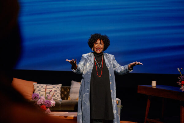 A female-presenting person gestures toward an unseen audience with arms extended out and palms up on a stage with a blue backdrop