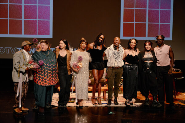 Several members of Open Television gather on a stage with two large faux windows behind them