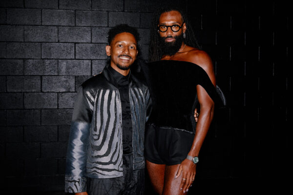 Two people with dark skin pose against a black brick wall
