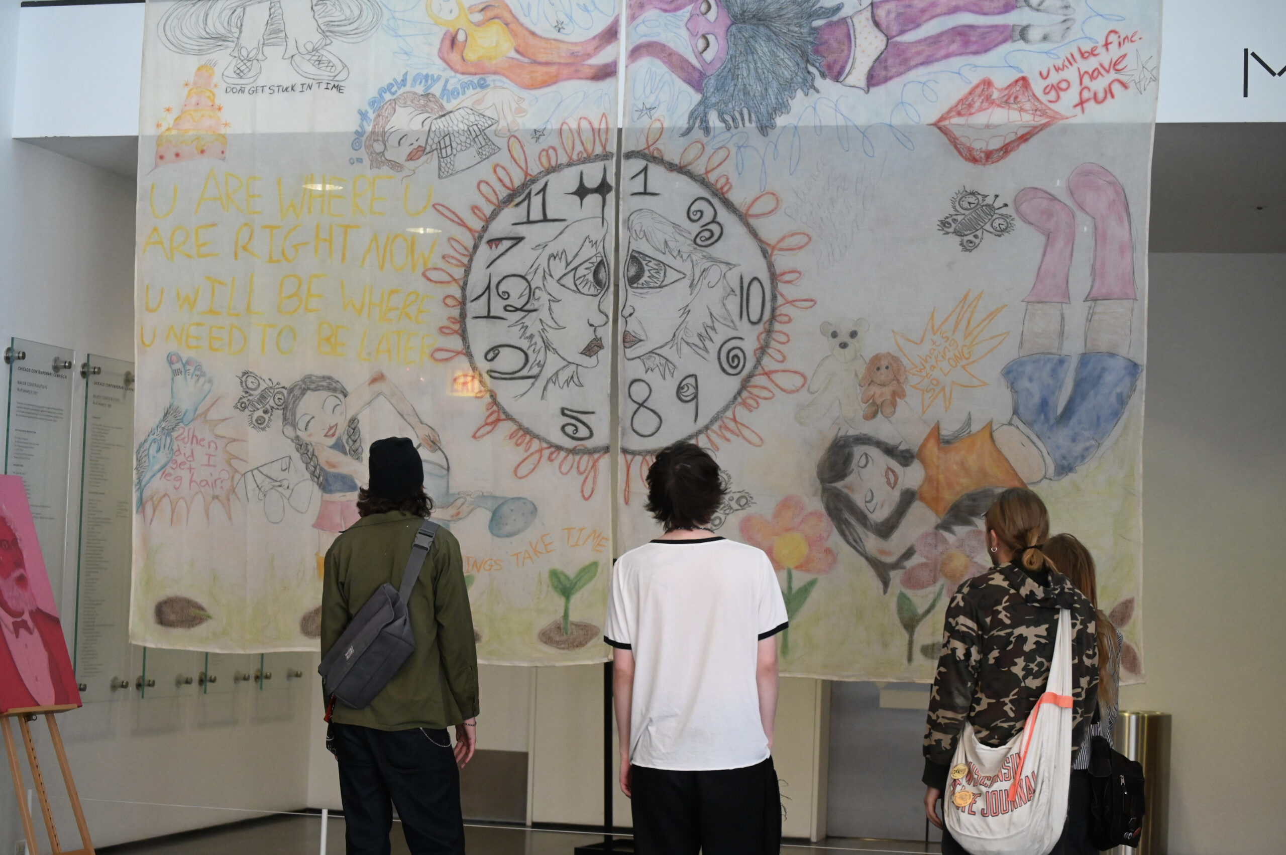 Three people stand in front of a large, colorful art piece displayed indoors. The artwork features a central clock surrounded by various whimsical illustrations and text. Bright colors and playful designs dominate the mural-like piece.