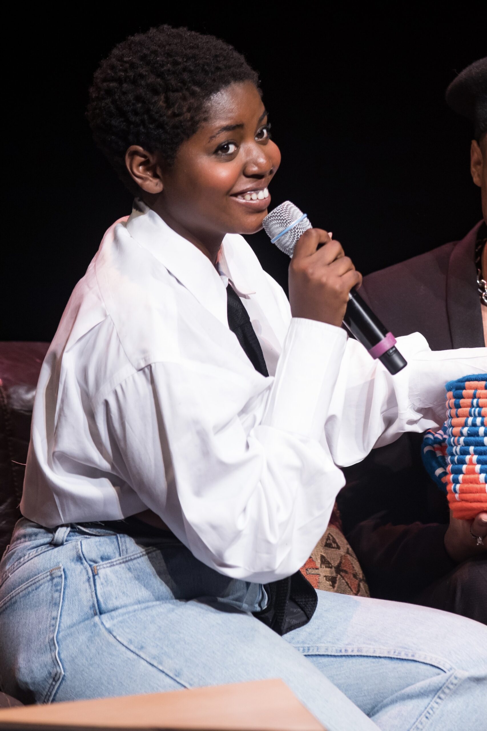 A female-presenting person with dark skin smiles over their right shoulder while holding a microphone up to their mouth