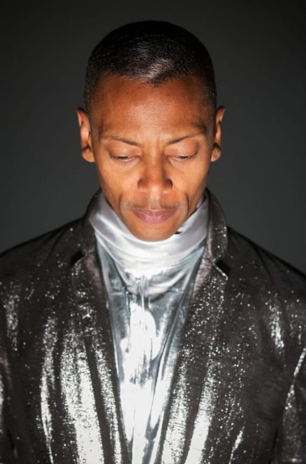 A person with caramel-toned skin looks down toward a light source that illuminates their face and highlights their shiny silver suit