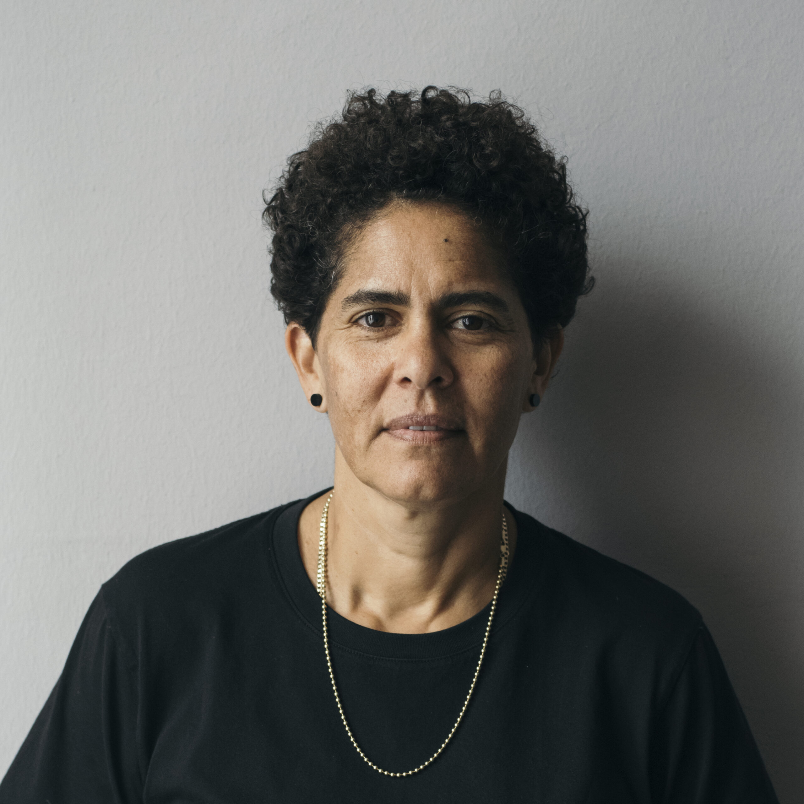 Portrait of a woman with short dark hair and olive skin, posing against a grey background with black clothing on