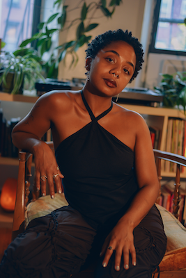 Portrait of a female-presenting person with medium-dark skin sitting in a vintage chair in a living room filled with plants and shelving