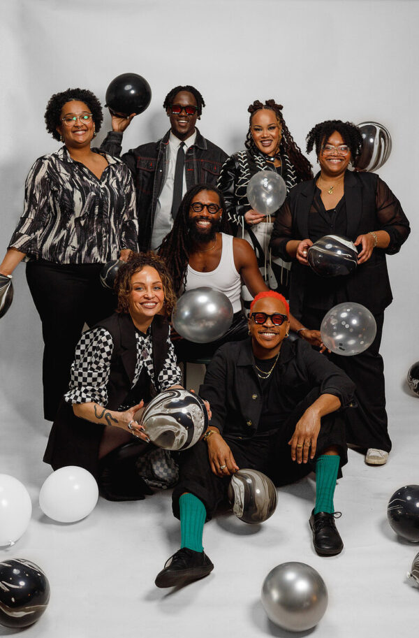 A large group of adults pose against a white backdrop with white, black, and silver ballons on the ground in front of them