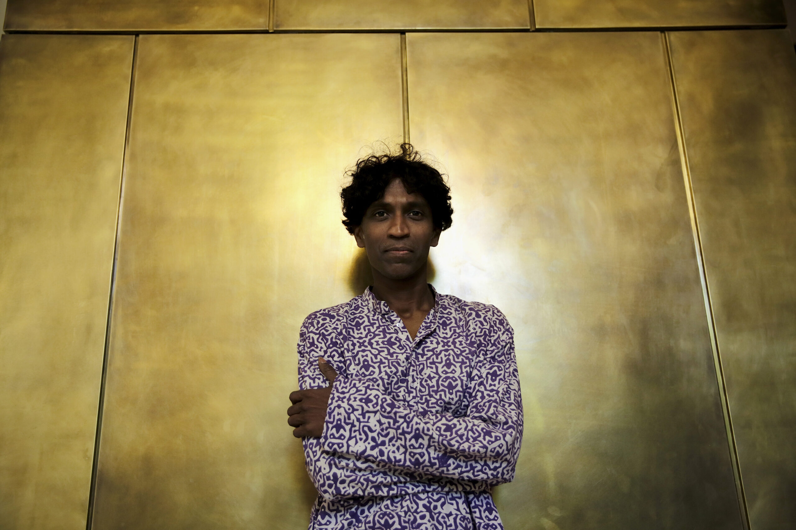 Portrait of a dark-skinned person with curly hair against large, shiny gold tiles