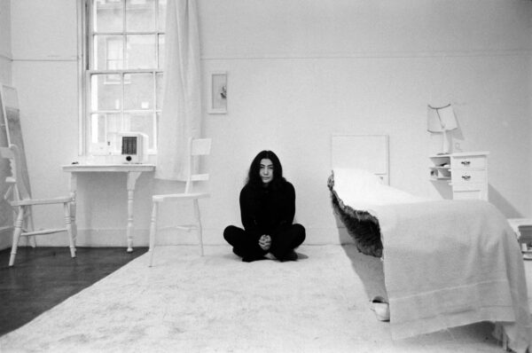 Black and white photo of Yoko Ono sitting crossed legged in a room where everything, from the bed to the artwork hanging on the wall, is a half slice of the object