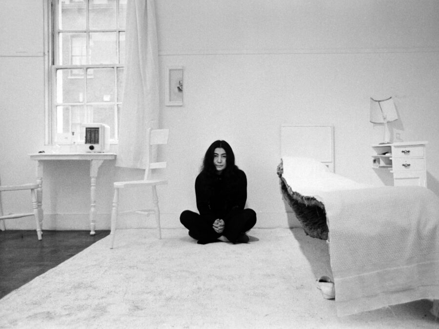 Black and white photo of Yoko Ono sitting crossed legged in a room where everything, from the bed to the artwork hanging on the wall, is a half slice of the object