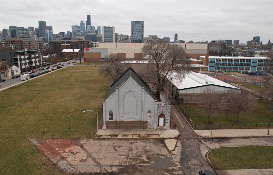 An old grey church with bricked-up windows among several empty lots and a warehouse. In the background is a Target store; a cityscape lies beyond that.