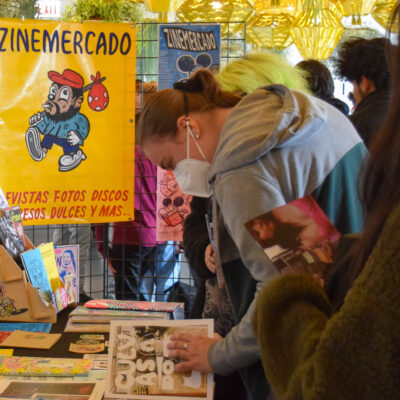 Several people browse a table filled with zines near a sign that reads Zine Mercadeo in a bright room with hanging planters