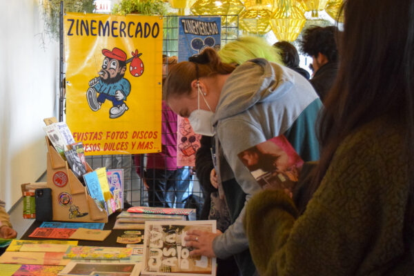 Several people browse a table filled with zines near a sign that reads Zine Mercadeo in a bright room with hanging planters
