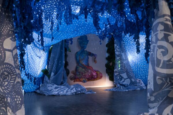 An art gallery features a dynamic installation with blue and white draped fabrics creating a tent-like space around a painting cast in shadow of a colorful seated figure