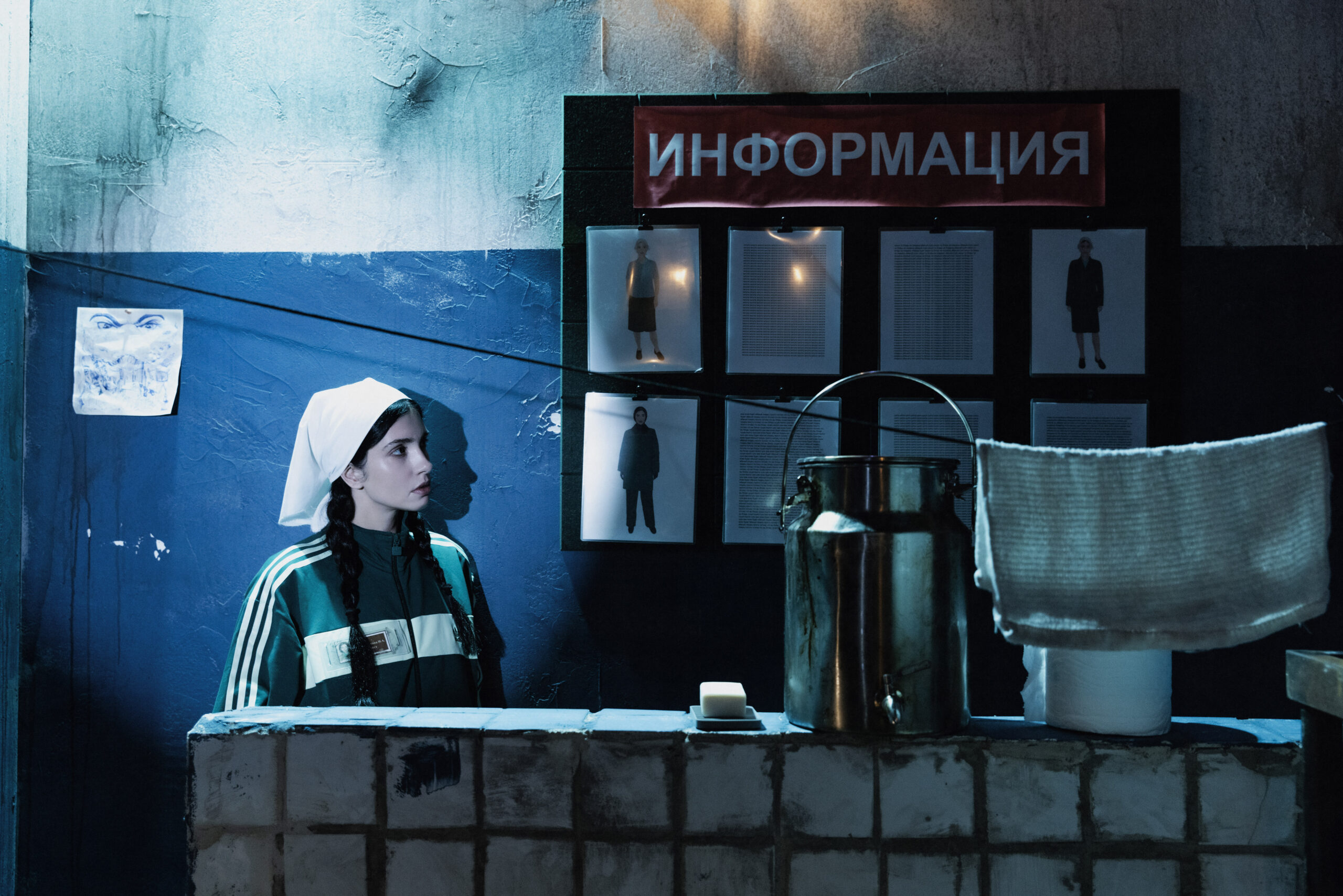 A spotlight shines on a person sitting behind a tiled half wall next to a bulletin board with pictures and a headline in Russian on it.