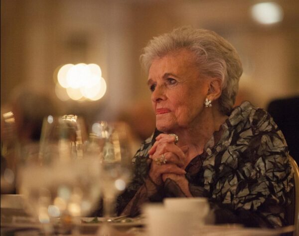 A woman with gray hair sitting at a table filled with wine glasses and coffee cups clasps her hands together under her chin while looking off into the distance. A warm glow from the left highlights her face.