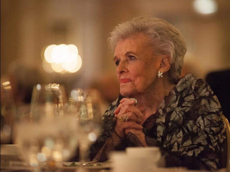 A woman with gray hair sitting at a table filled with wine glasses and coffee cups clasps her hands together under her chin while looking off into the distance. A warm glow from the left highlights her face.
