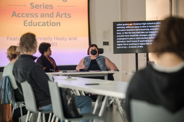 A group of people sit around tables formed into an l-shape in front of a screen projecting an orange slide with text about access and arts education with a tv to the right displaying a long paragraph of text