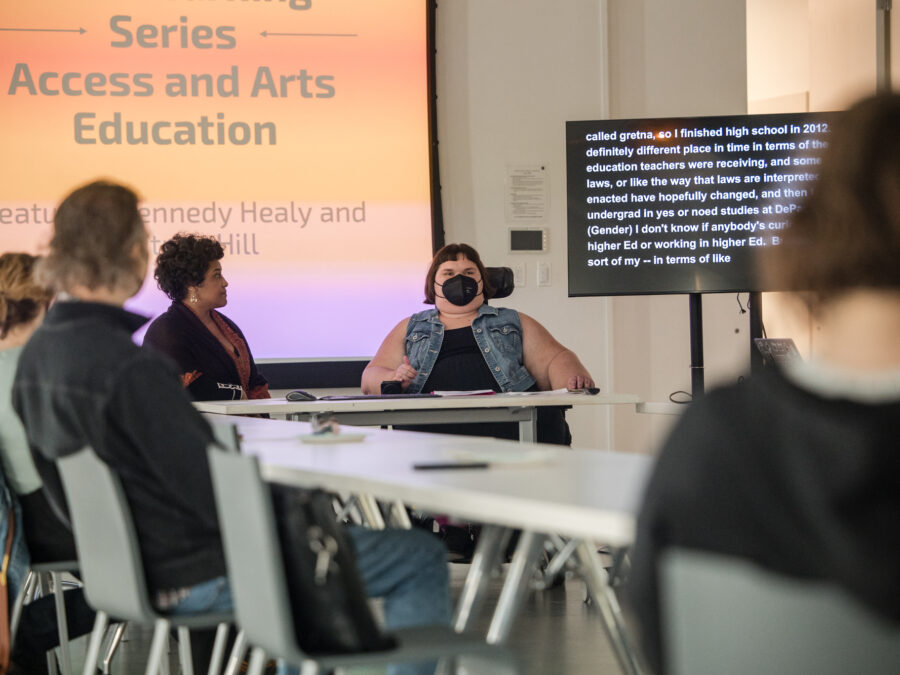 A group of people sit around tables formed into an l-shape in front of a screen projecting an orange slide with text about access and arts education with a tv to the right displaying a long paragraph of text