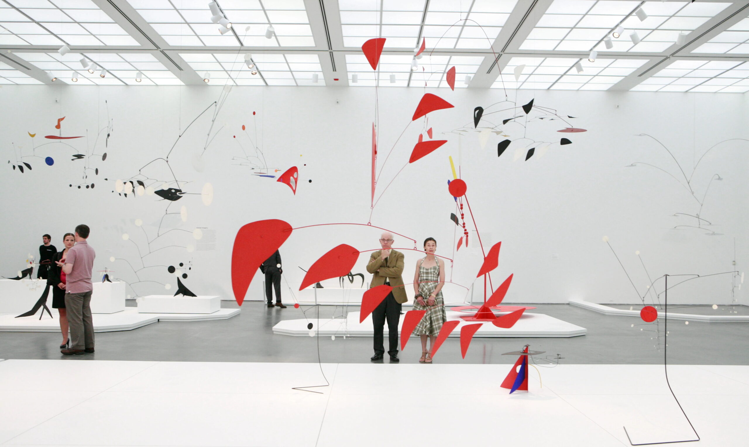 People looking at art in a brightly lit gallery featuring an extensive display of Alexander Calder's sculptures, characterized by their abstract shapes and vibrant colors. At the forefront is an burnt orange mobile suspended in air.