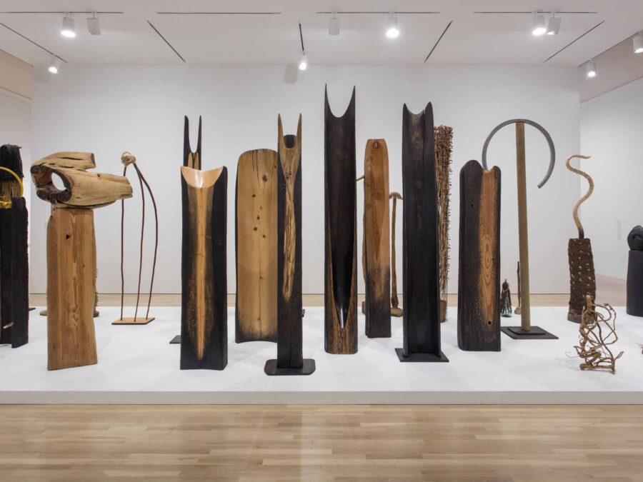 A grouping of abstract wooden sculptures, some carved into two points at the end, others with items attached to them, on a white platform in a well-lit gallery space