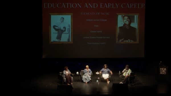 Four people sit on a stage in a row with a powerpoint presentation slide projected onto a large screen behind them