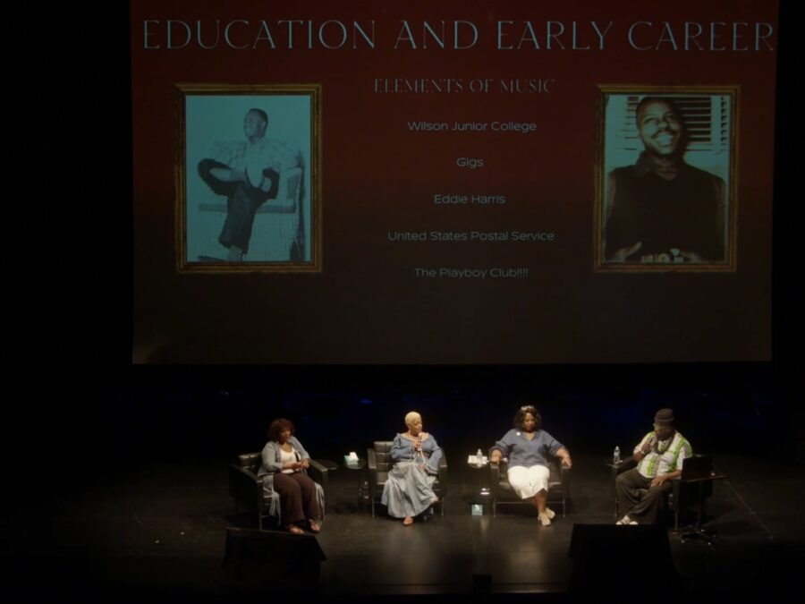 Four people sit on a stage in a row with a powerpoint presentation slide projected onto a large screen behind them